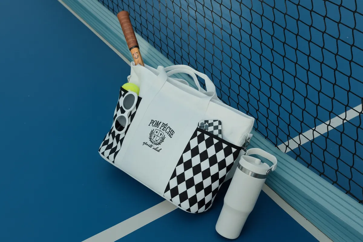 A white checkerboard tote bag with a pickleball racket sits on a blue court by the net.