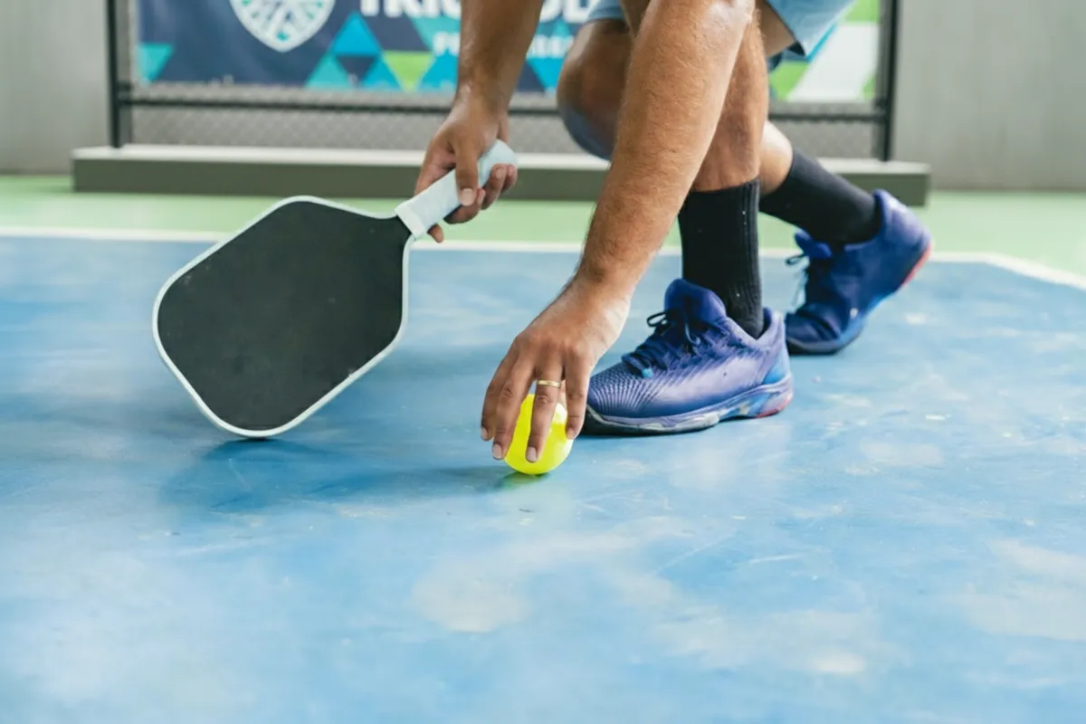 A pickleball player bends down to pick up a yellow ball from the blue court surface.