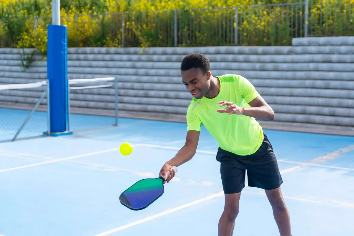 A young man wearing a bright yellow-green shirt plays pickleball on an outdoor blue court, leaning forward to hit a yellow ball with his paddle.