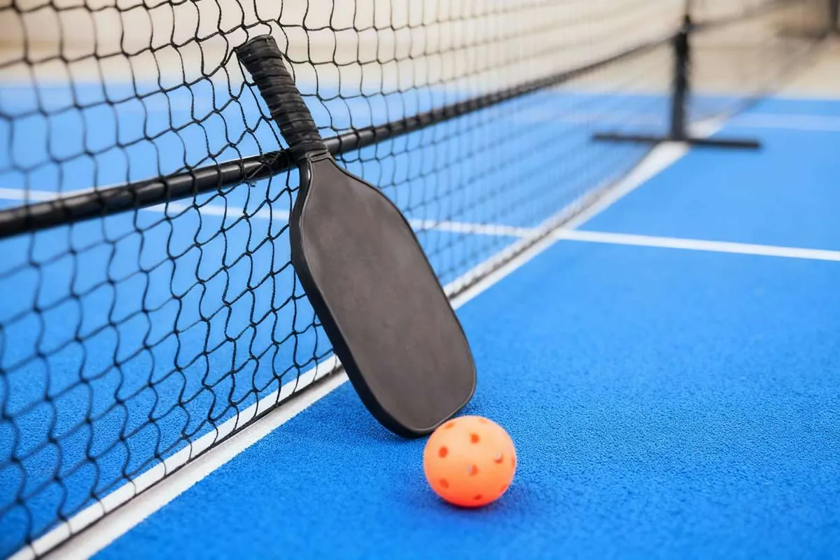 A black pickleball paddle leans against the net on a bright blue pickleball court, with an orange ball resting on the surface.
