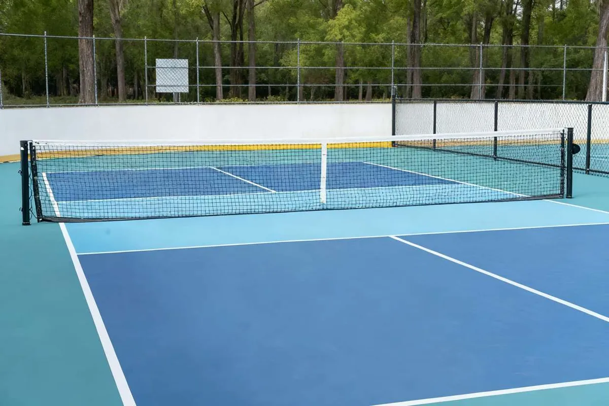 An empty outdoor pickleball court with a blue and green surface, white lines, and a net set up in front of a chain-link fence.