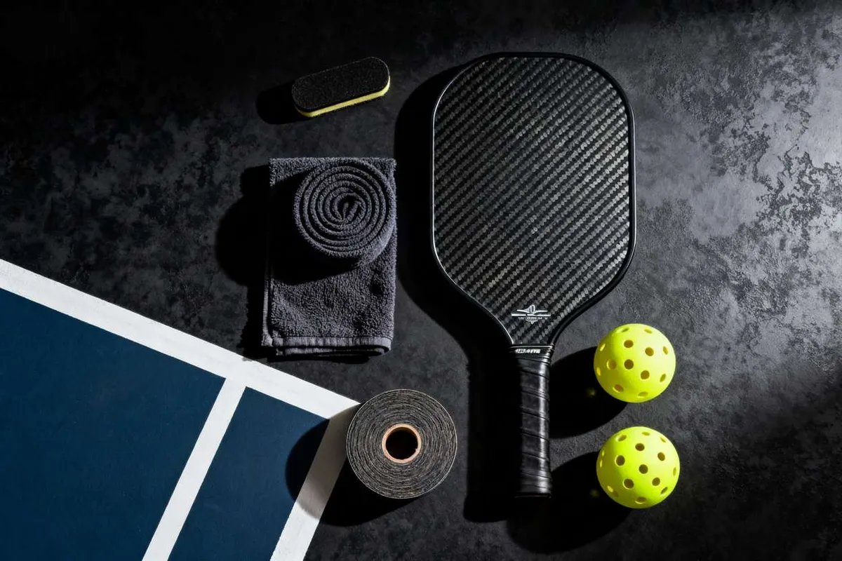 A flat lay of essential pickleball gear, including a carbon fiber paddle, two yellow balls, a towel, grip tape, and a paddle eraser, arranged on a dark court.
