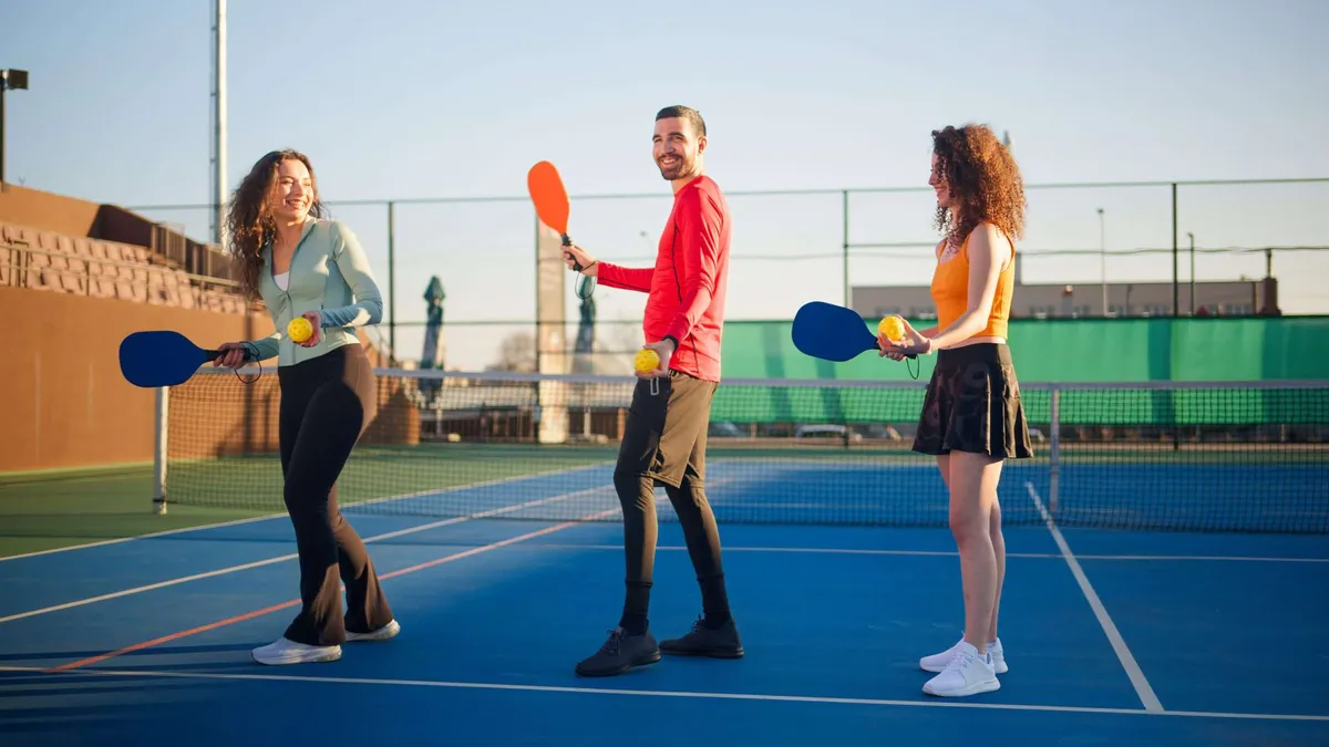 A group of three happy friends smiling and holding paddles on a blue pickleball court during a sunny afternoon.