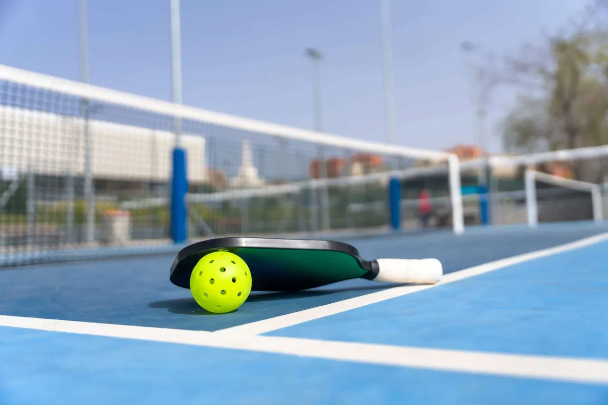 A green pickleball paddle and a yellow pickleball on a blue court with the net in the background.