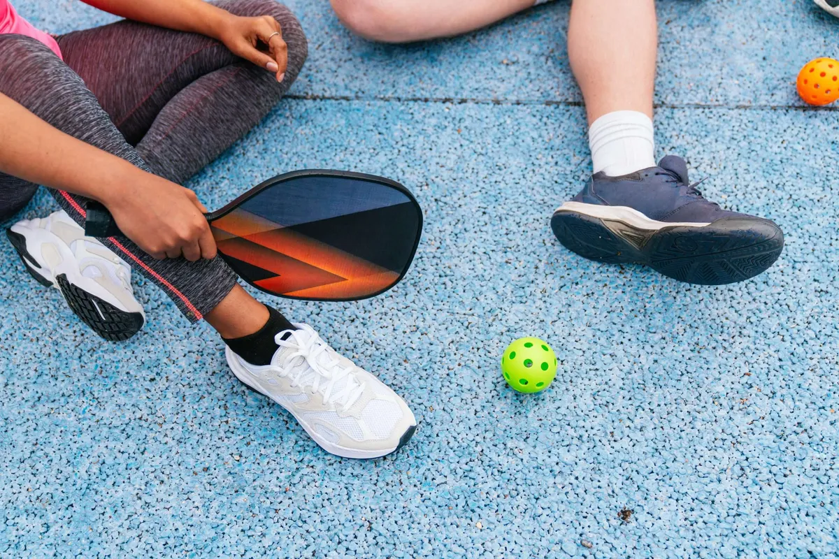 Two people sit on a court with a pickleball paddle and a green ball between them.