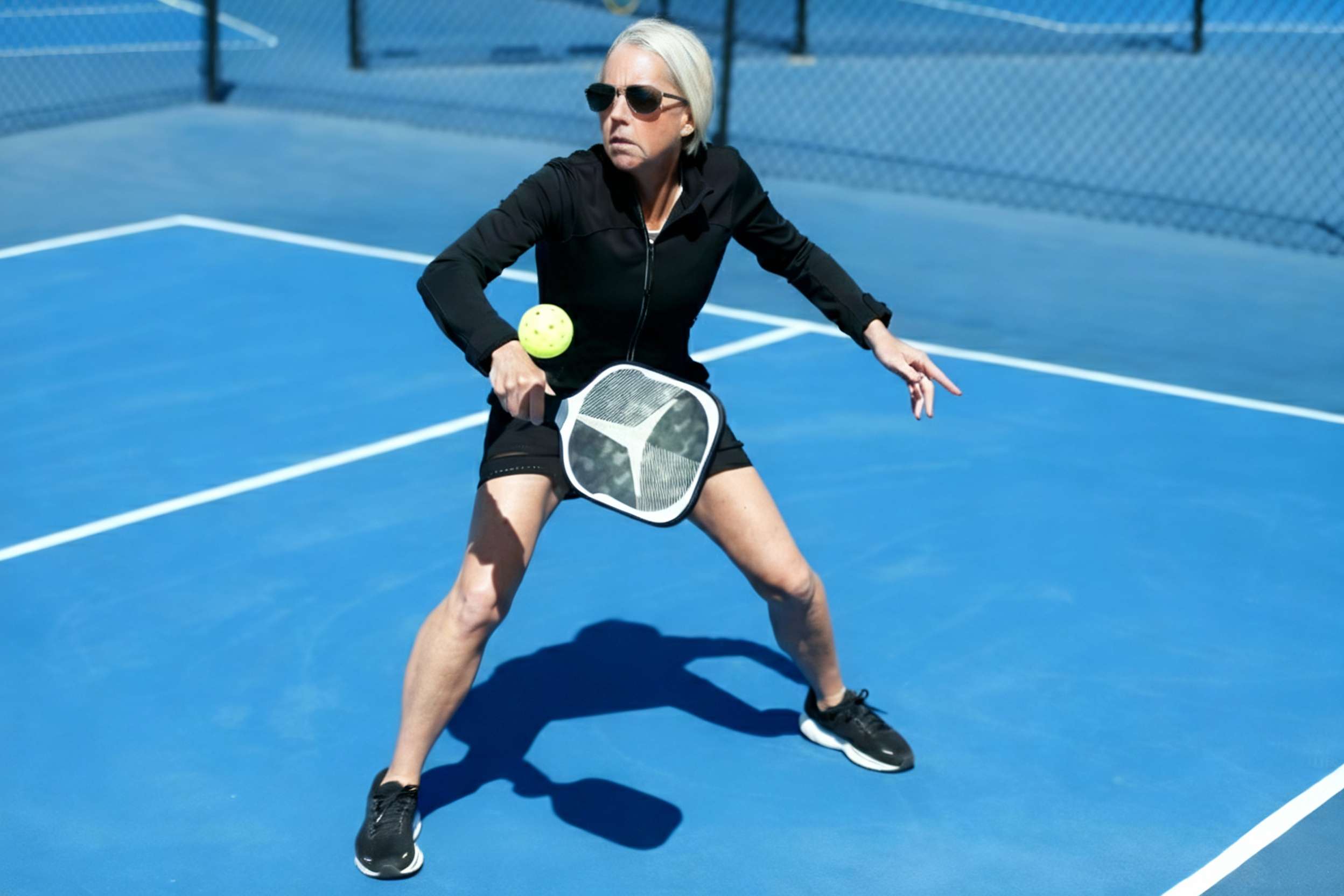 A female player demonstrates proper pickleball backhand technique with a focused athletic stance as she makes contact with the ball on a blue court. 