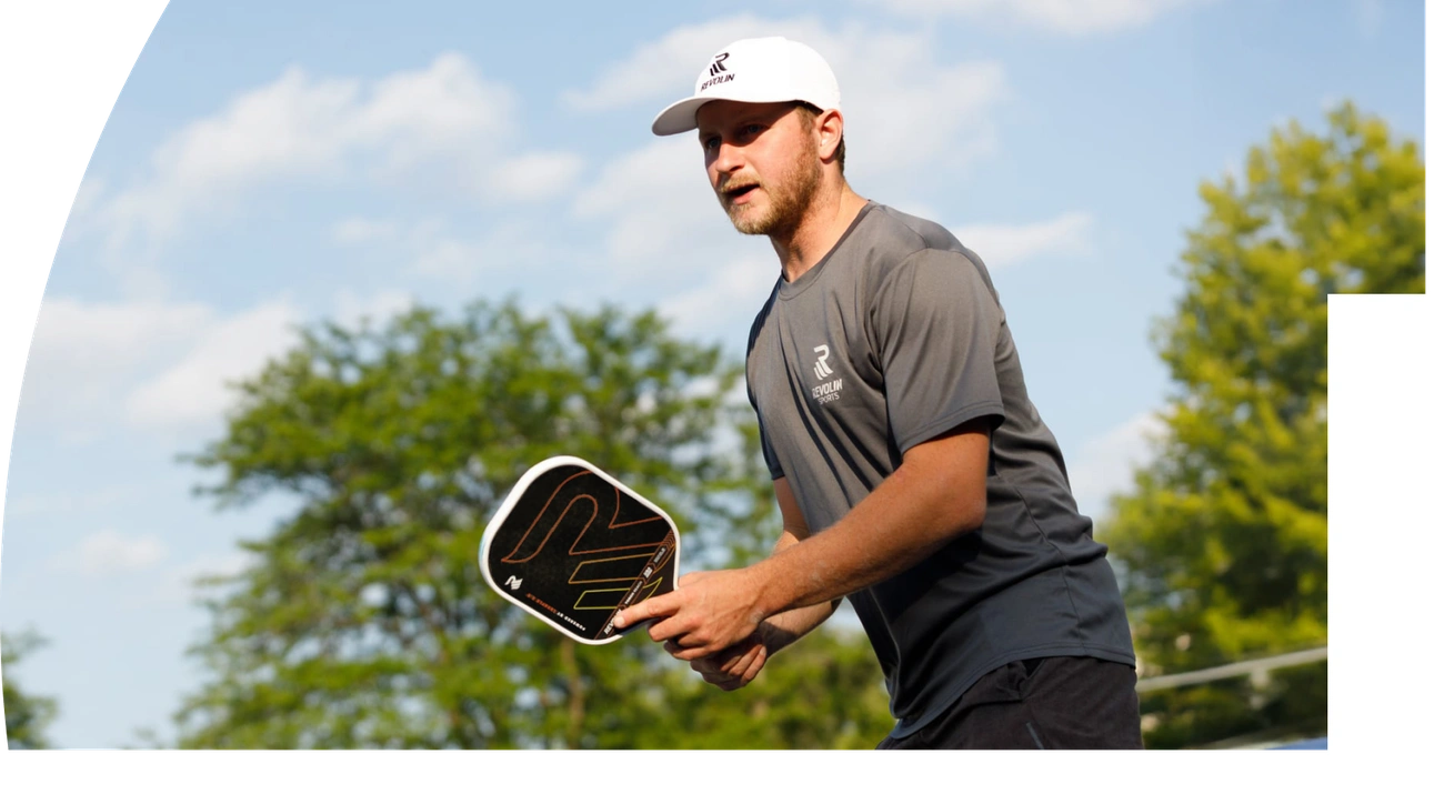 A focused male pickleball player in a grey shirt assumes the proper ready stance, demonstrating a key technique on an outdoor court.