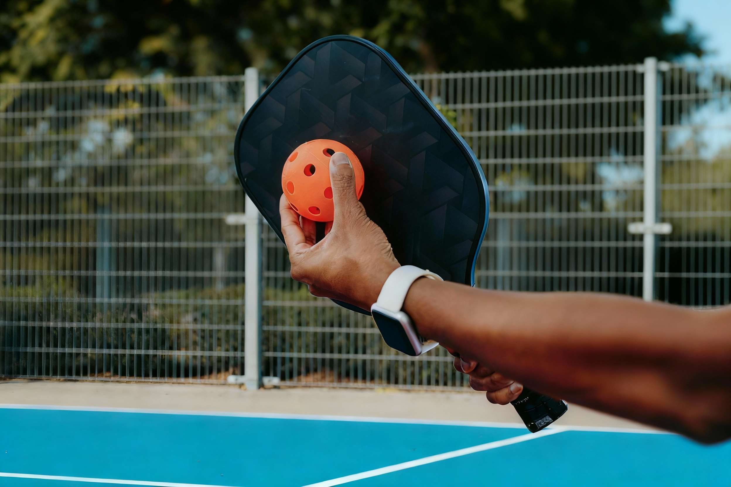 A close-up of a hand holding an orange pickleball against a paddle, ready to serve