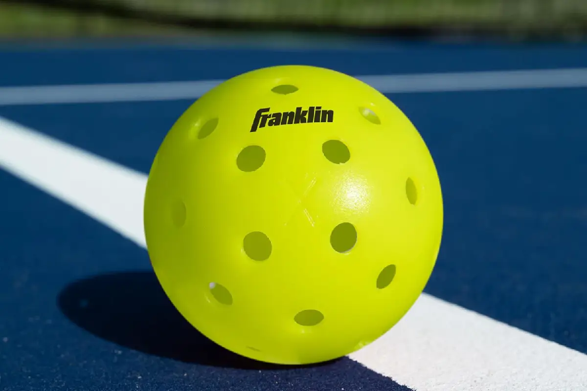 A yellow Franklin outdoor pickleball, a popular choice for the best pickleball balls, resting on the white line of a blue court.