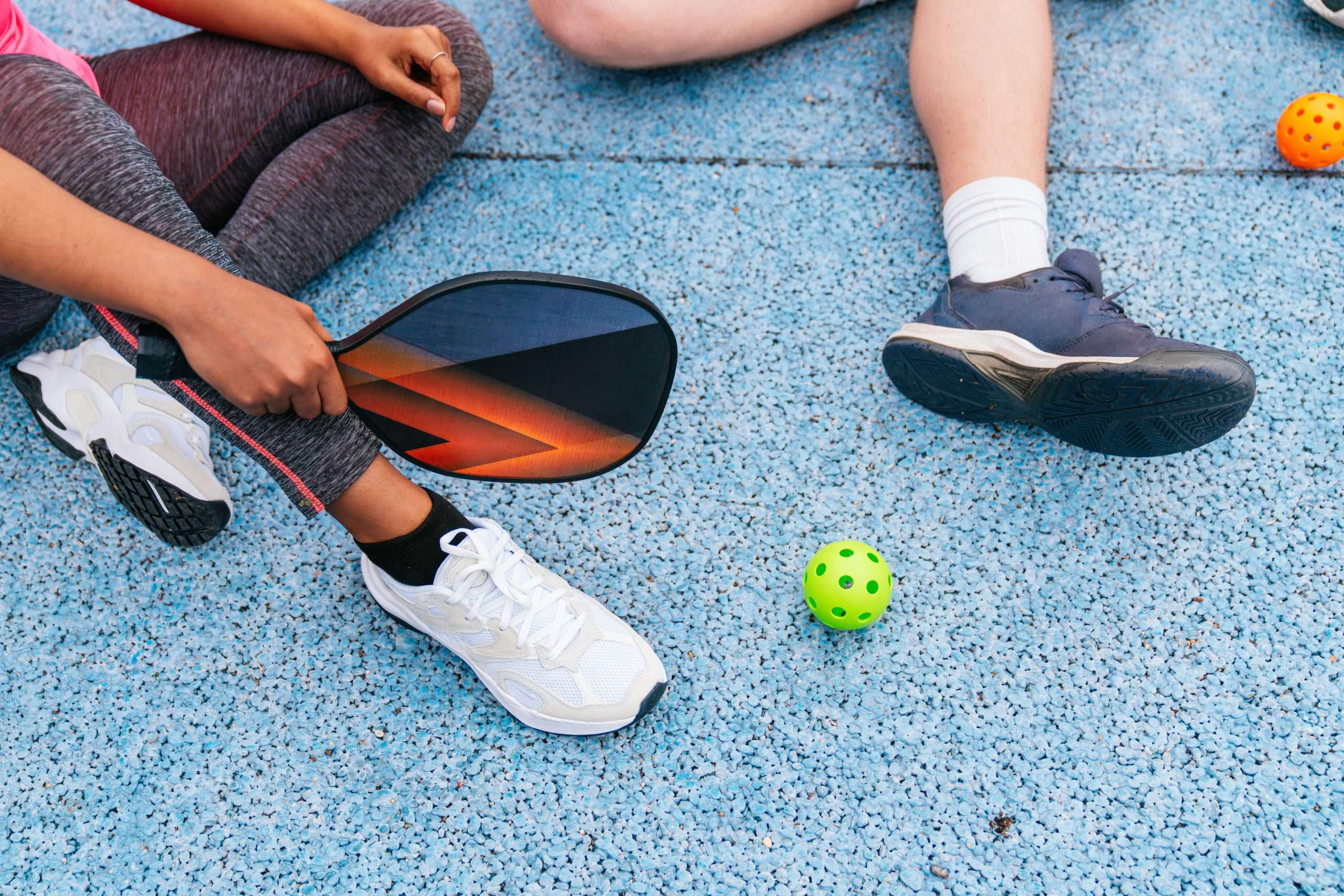 Two people sit on a court with a pickleball paddle and a green ball between them.