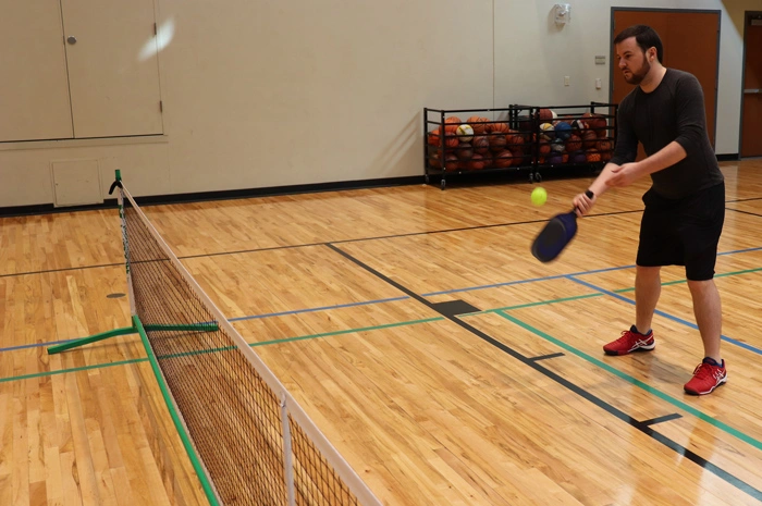 A man practices his pickleball forehand on a temporary gymnasium court, demonstrating how to play indoors with a portable net. 