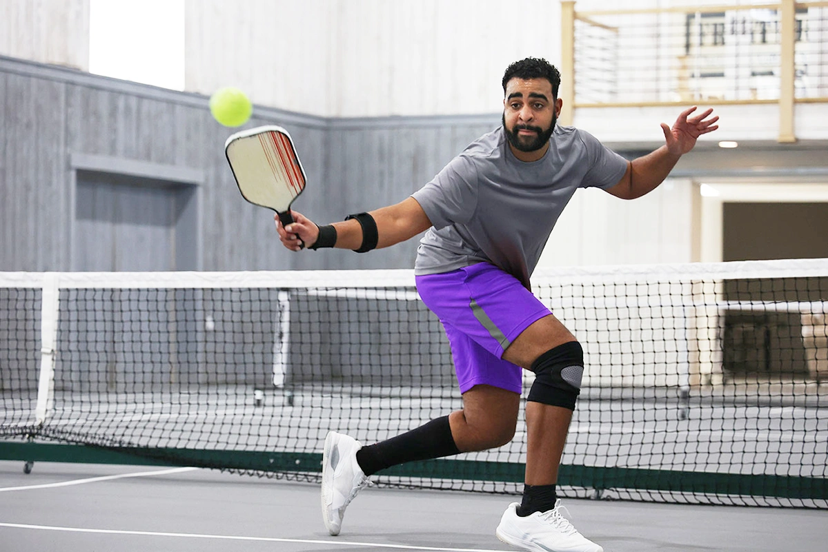 A male pickleball player demonstrating correct backhand technique with a strong, athletic stance while hitting a volley on an indoor court. 