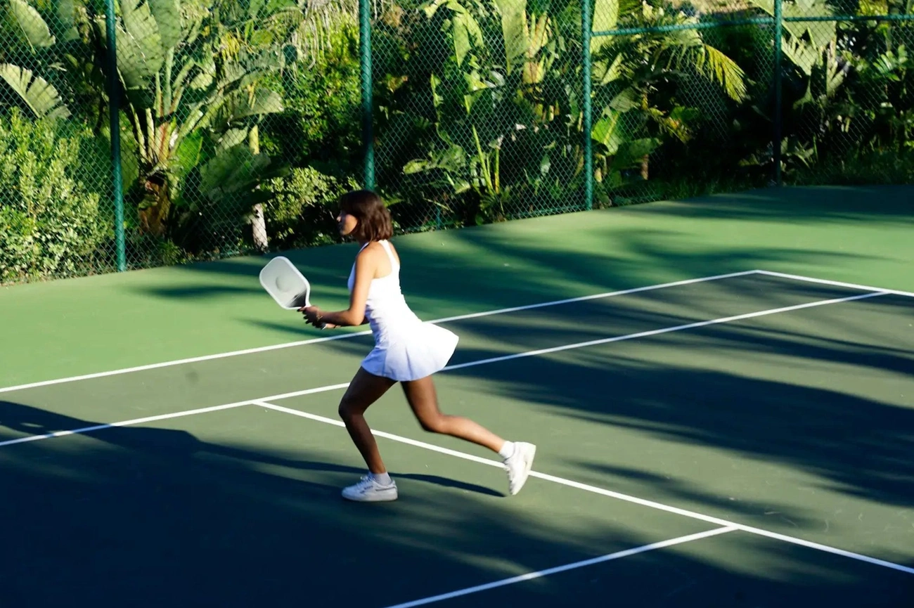 A woman in an all-white outfit hits a backhand shot on a lush, green tennis court.