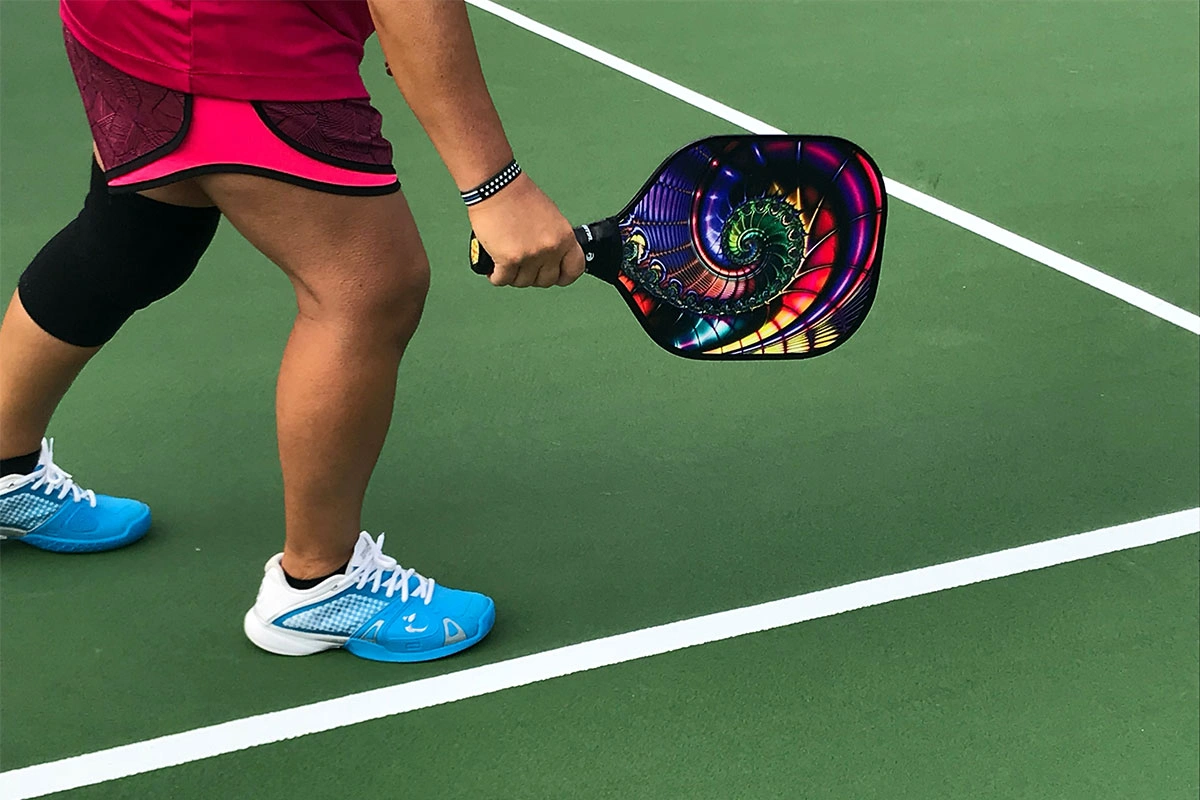 Player holding a colorful pickleball paddle on a green court wearing blue and white shoes and a black knee brace.