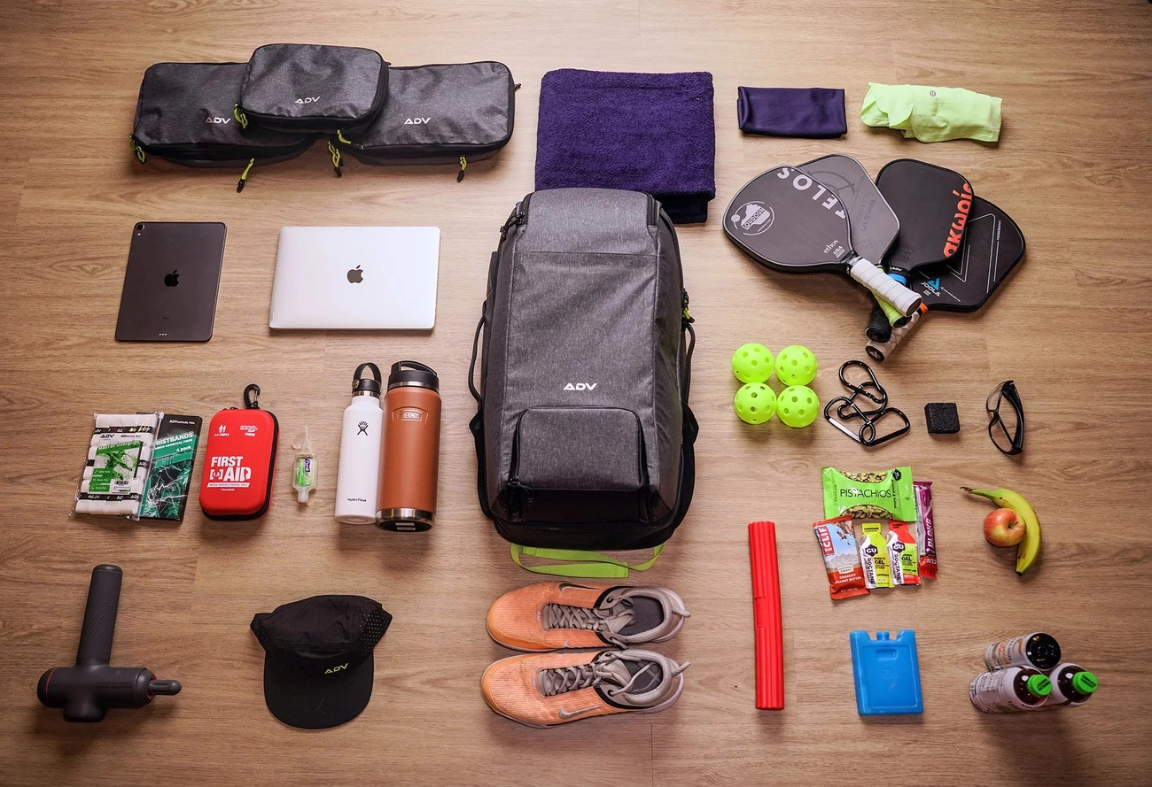 A flat lay of pickleball gear packed in and around a grey ADV backpack, including paddles, balls, shoes, and tech on a wood floor.