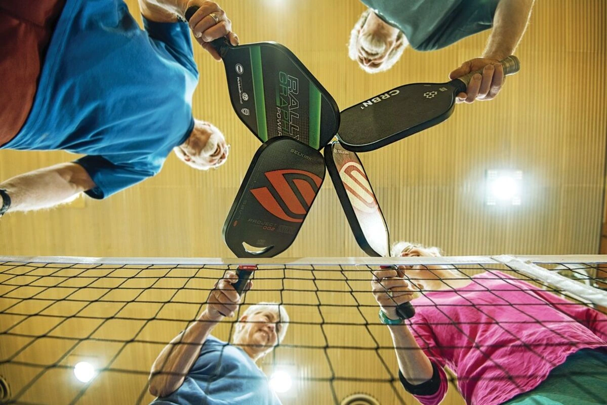 A low-angle view from under the pickleball net showing four players touching their paddles together in a team huddle on an indoor court.
