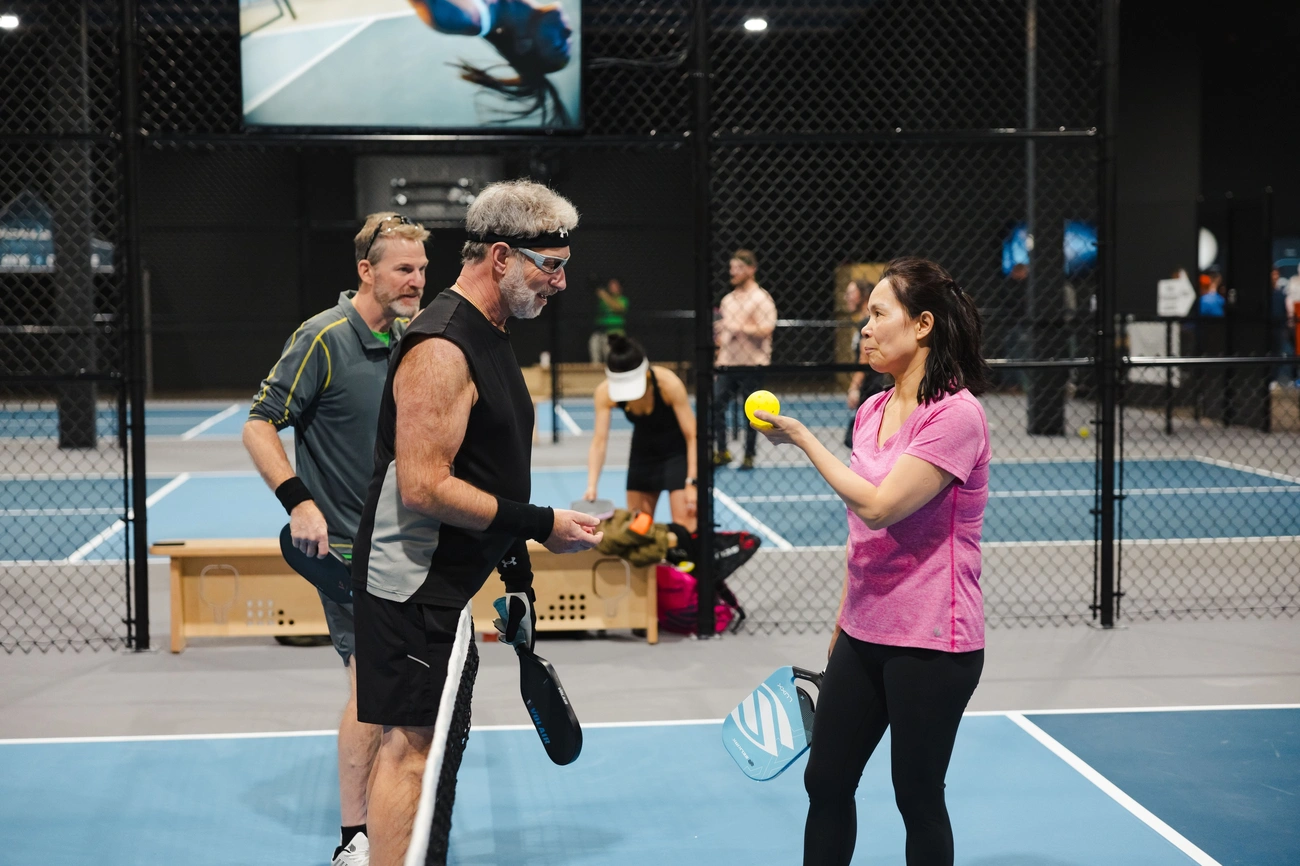 A woman in a pink shirt holds a yellow pickleball while talking to two male players on a blue indoor pickleball court.