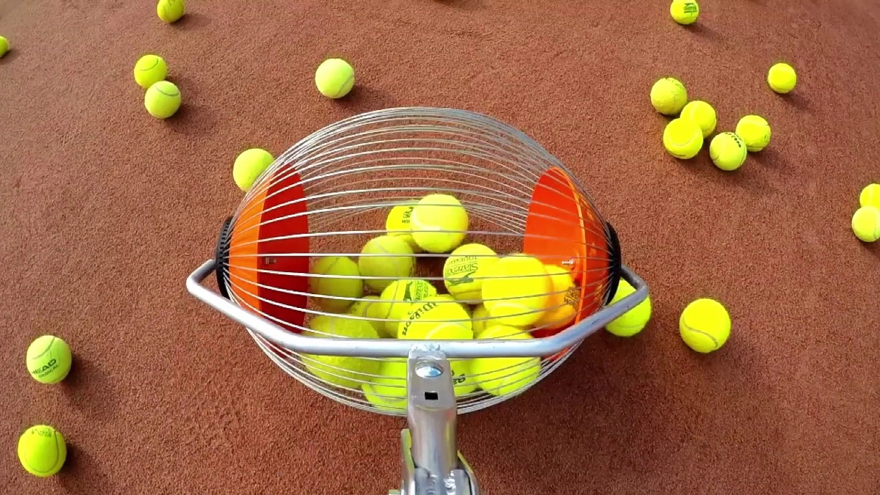 A rolling tennis ball hopper partially filled with yellow balls, sitting on a reddish-brown tennis court with many other balls scattered around it.