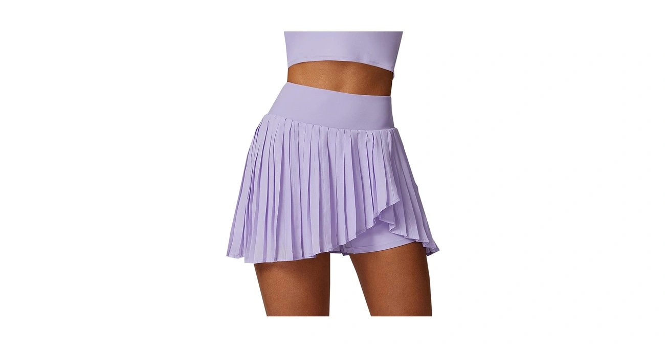 A woman modeling a high-waisted, lavender purple pleated athletic skirt with an asymmetrical hem, shown isolated on a white background.