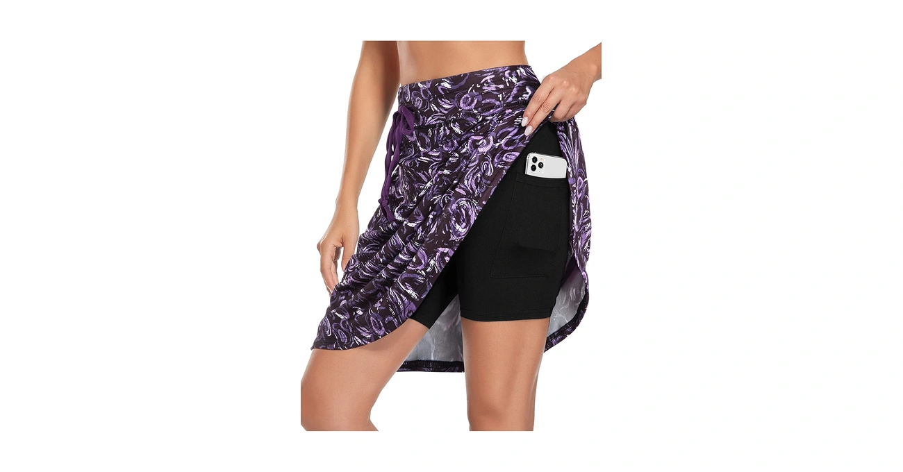 A woman wearing a purple patterned pickleball skort lifts the skirt layer to reveal a smartphone in the side pocket of the black built-in shorts.