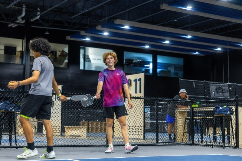 Two young men playing pickleball on a blue indoor court, one smiling at the camera while holding his paddle.